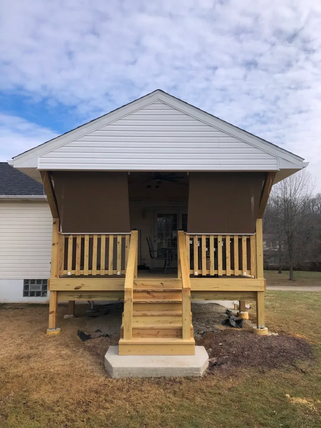Wooden porch with brown shades, steps, and white house siding. Cloudy sky.