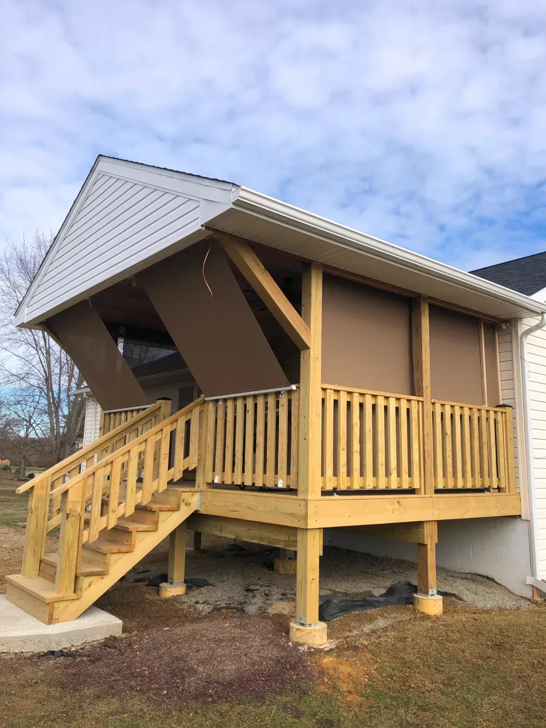 Wooden deck with steps, enclosed with brown shades, attached to a white-sided house under a cloudy sky.