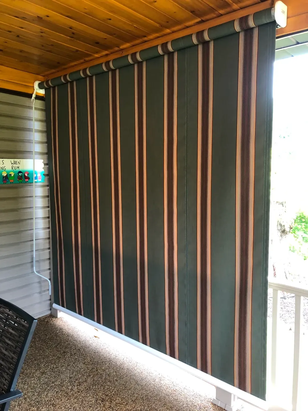 Green and brown striped outdoor roller shade, partially extended, on a porch.
