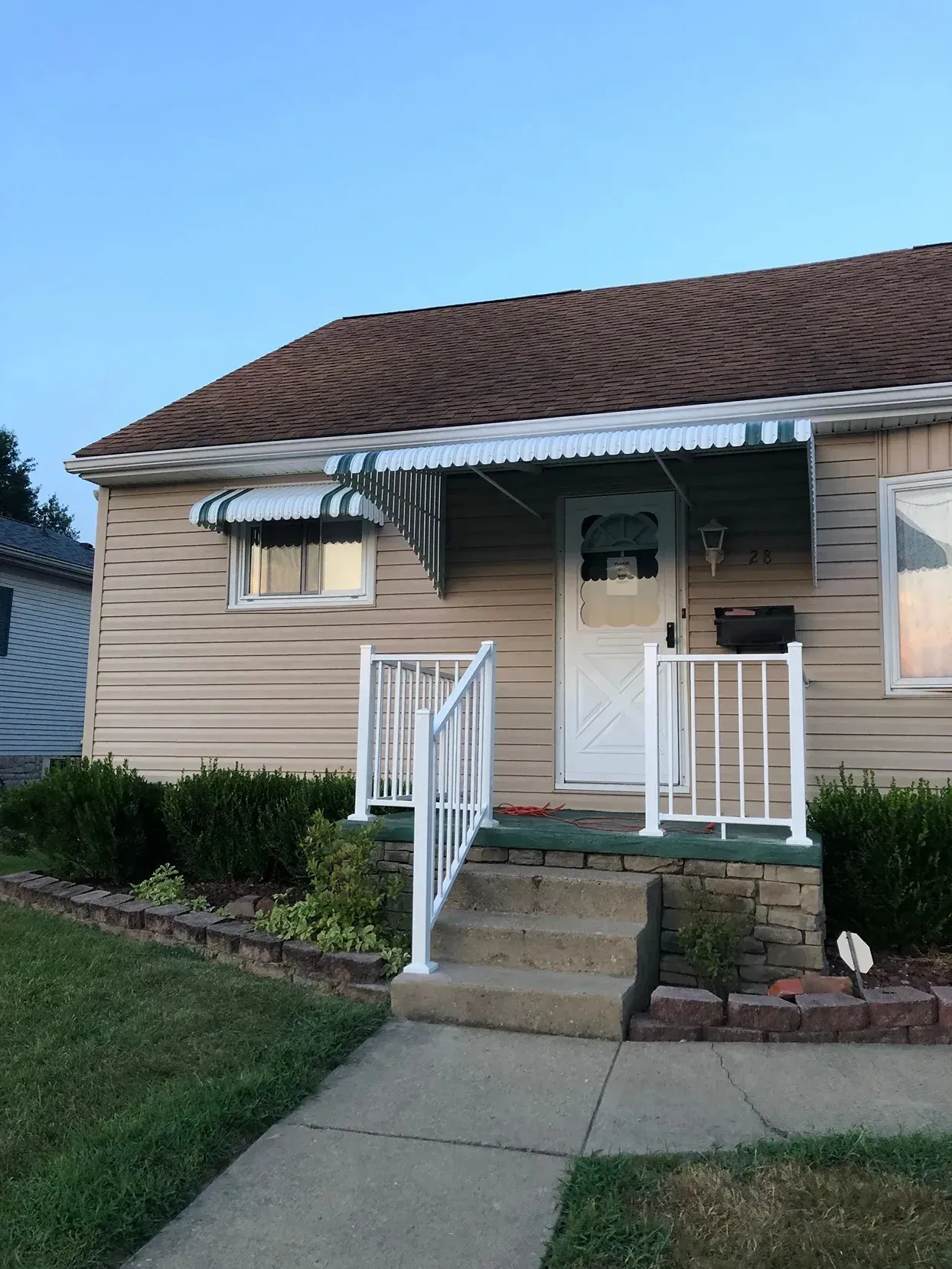 Tan house with brown roof, green and white awnings over door and window. White railing and steps to front door.