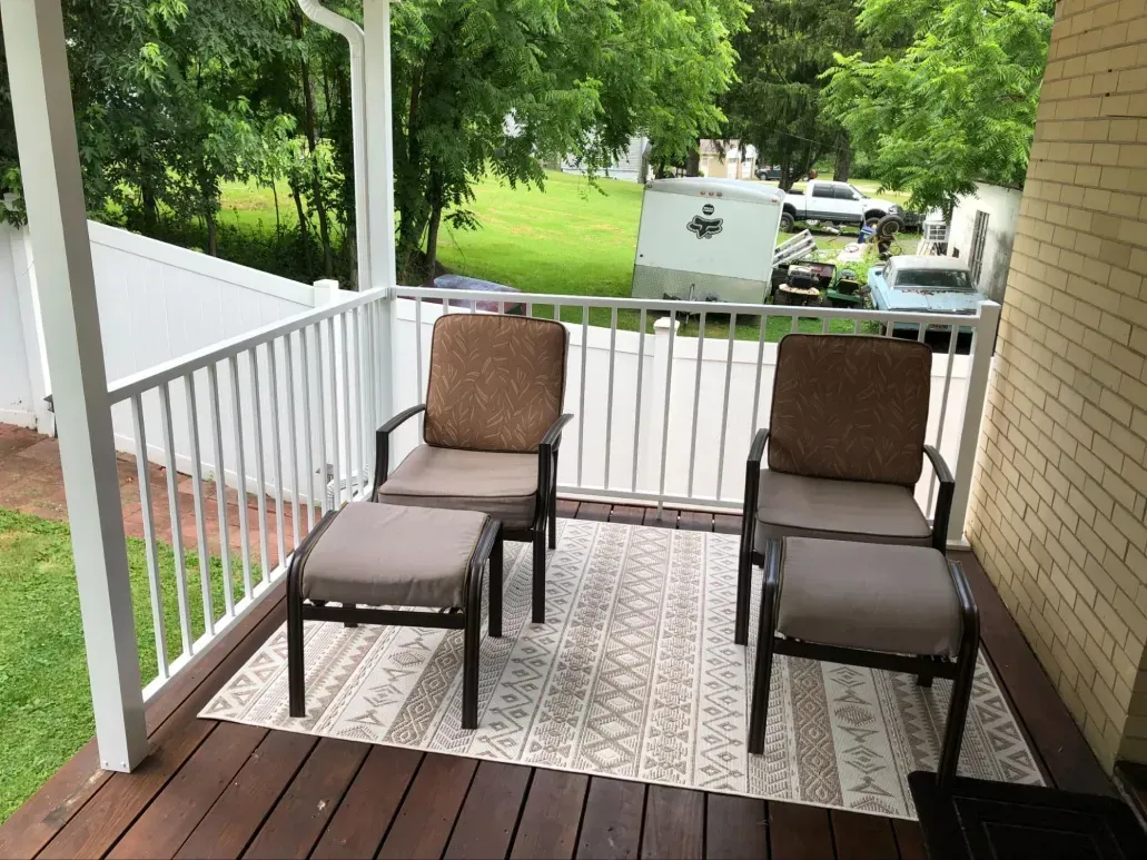 Two chairs and ottomans on a porch with a rug, looking out at a street with cars.