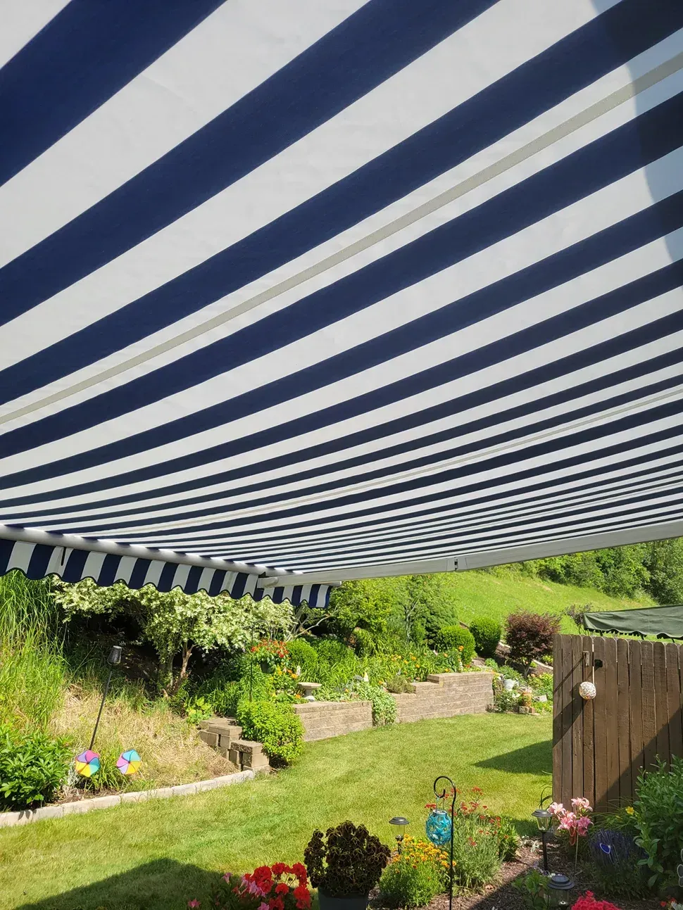 Blue and white striped awning shades a green backyard with flowers and stone retaining walls.