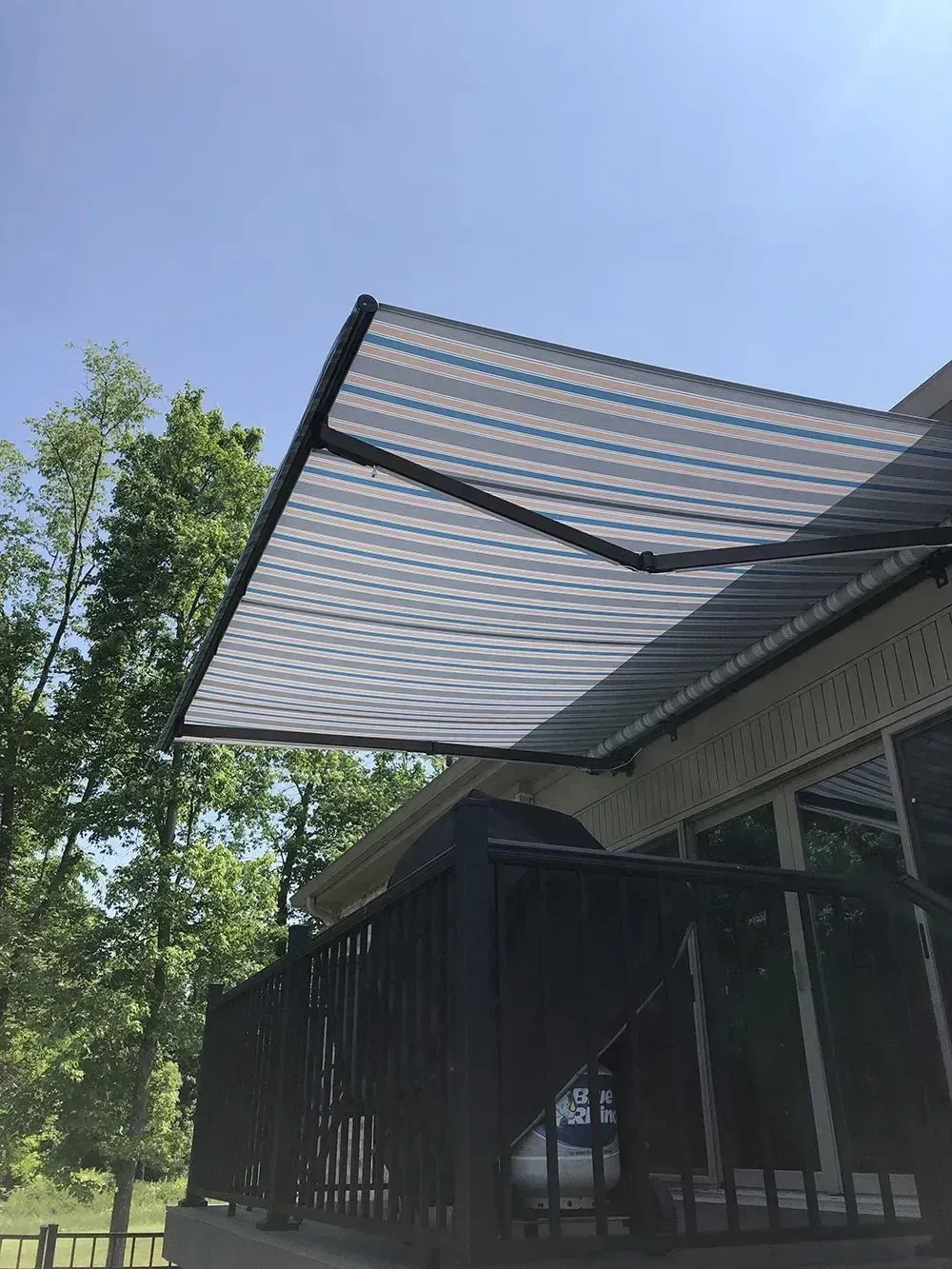 A retractable striped awning extends over a deck. The sky is blue, trees are in the background.