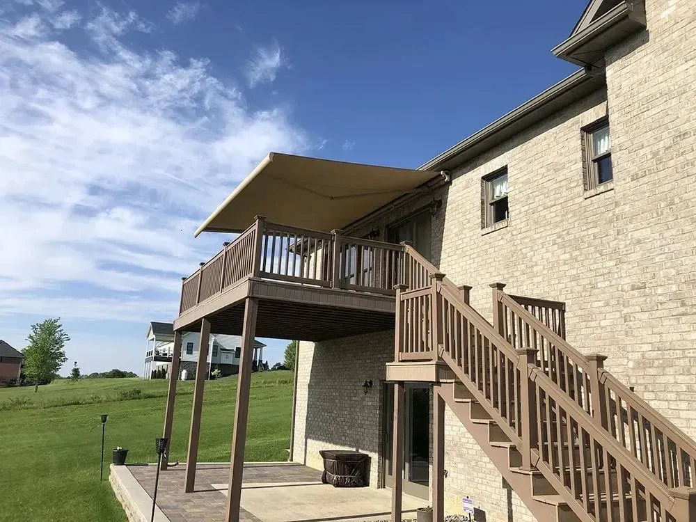 Tan retractable awning over a wooden deck attached to a brick house. Sunny day.
