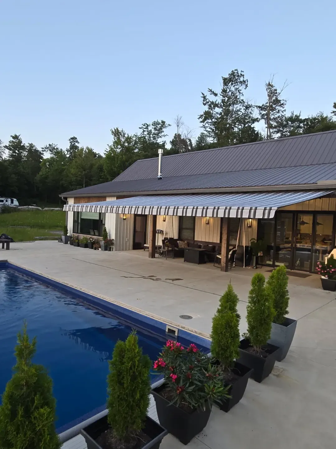 Poolside patio with blue and white awning. Trees in pots line the pool edge, house in background.