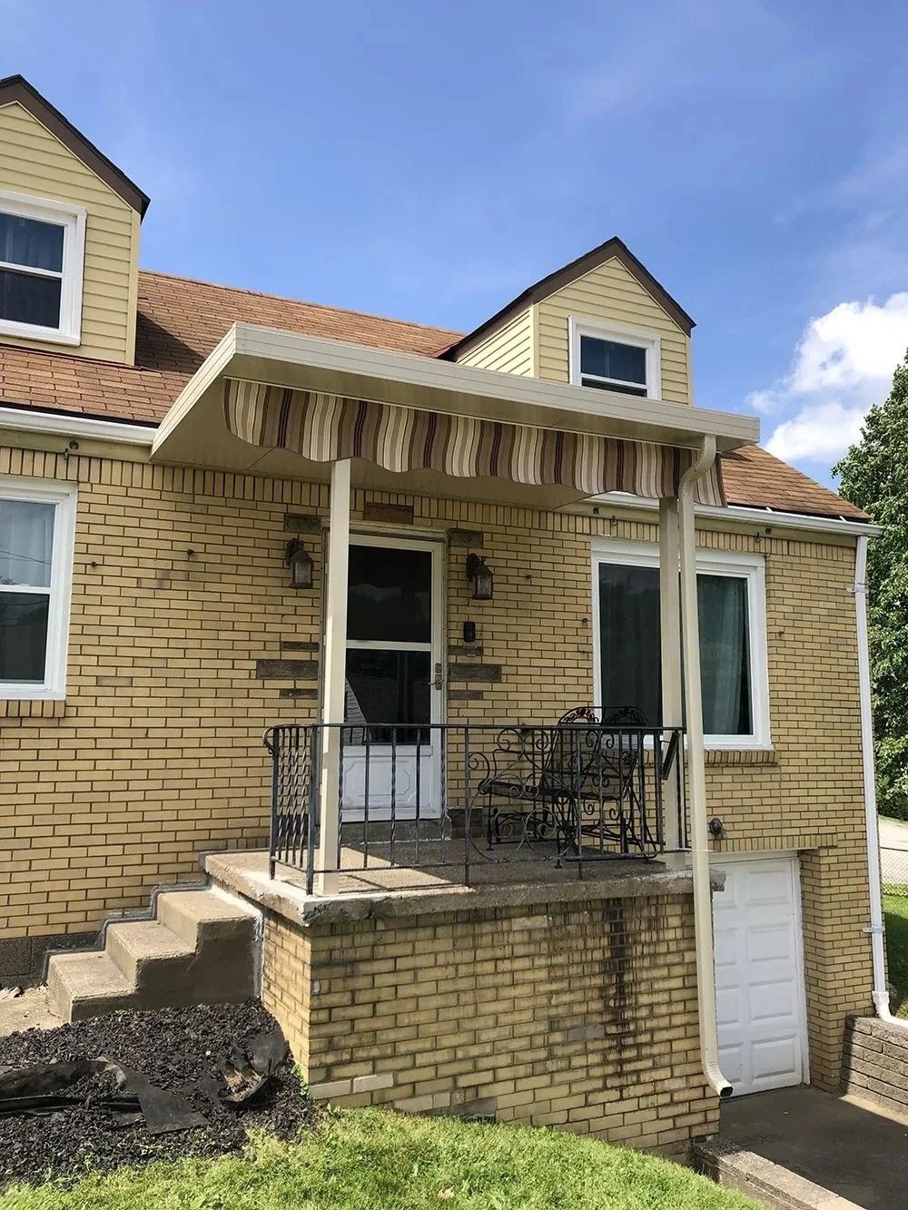 Yellow brick house with a porch and awning.  Steps lead up to a black railing and front door.