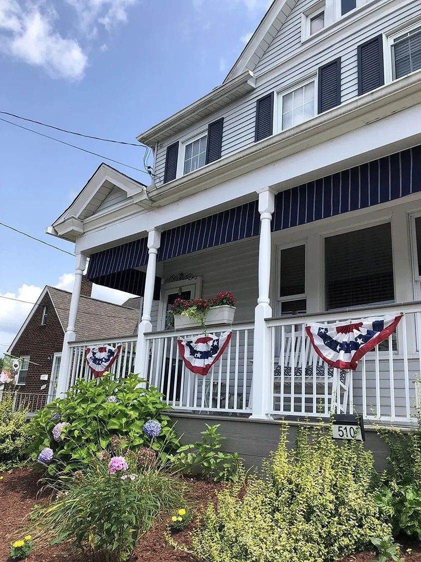 Two-story house with white porch, blue and white striped awnings, and red, white, and blue decorations.
