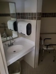 Accessible public restroom with sink, mirror, paper towel dispenser, and a wheelchair-accessible chair.