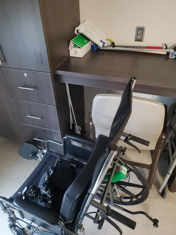 Wheelchair, chair, and desk in a room. Cabinet on the left, medical supplies on the desk.