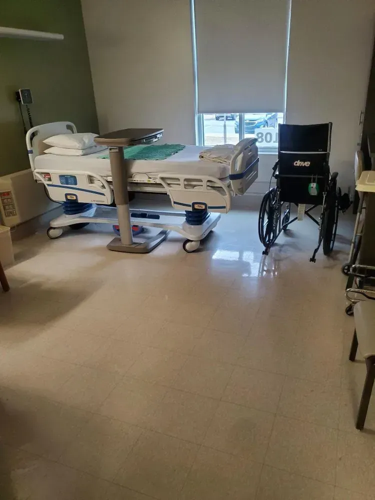 Hospital room with bed, tray table, and wheelchair. Bright room, light-colored floor, window with a blind.