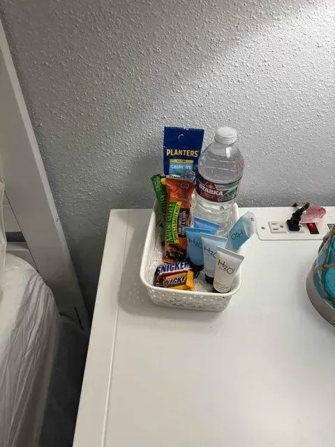A white basket on a white table holding snacks, a water bottle, and toiletries against a textured gray wall.