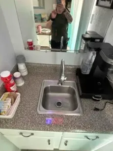 A woman in a mirror above a small sink with a coffee maker and supplies in a kitchen area.
