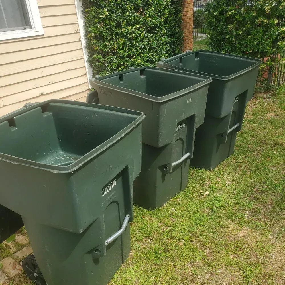Three green trash bins lined up on grass next to a building with a window.