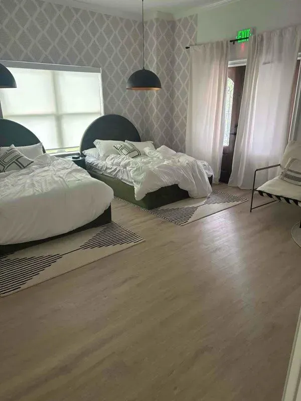 Bedroom with two beds, patterned wallpaper, light wood floors, and sheer curtains.