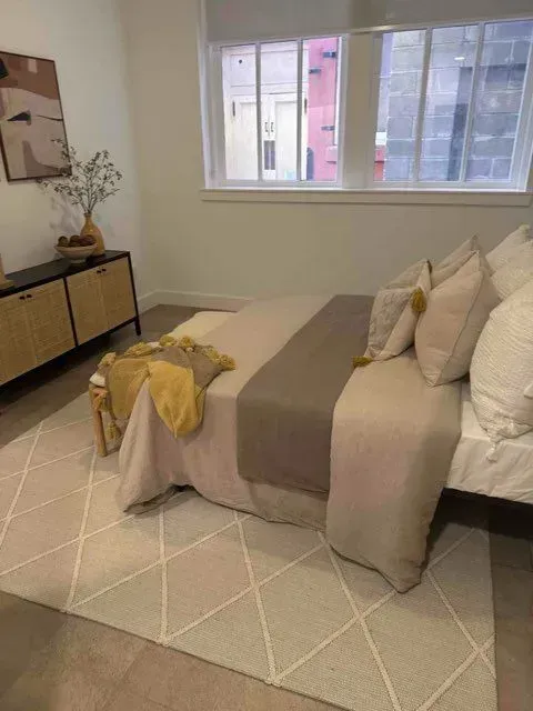 Bedroom with neutral tones. Bed has beige bedding, gray runner, & pillows. A patterned rug and a side table with decor are also present.