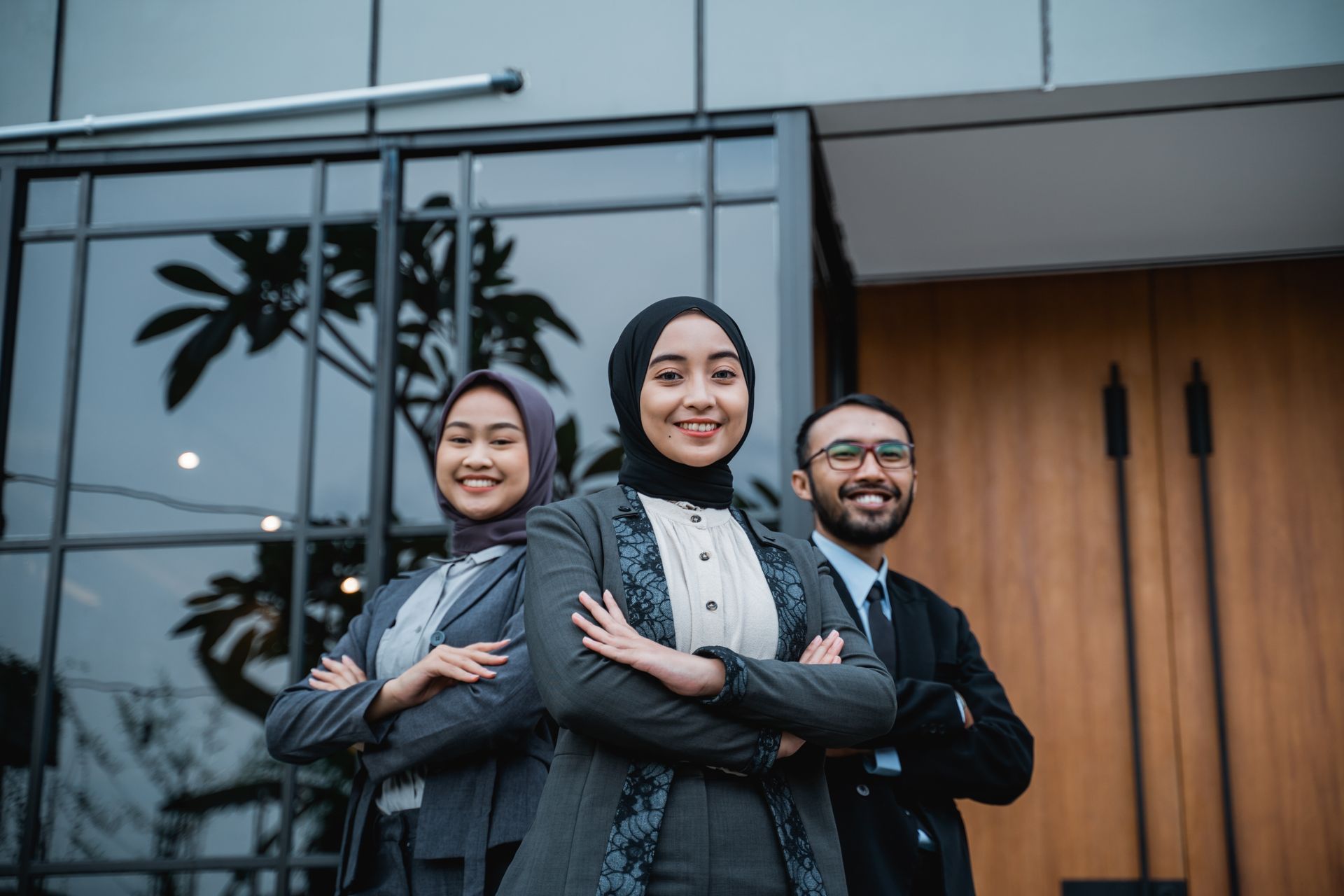 Three diverse Muslim Leaders smiling, arms crossed, standing in front of a modern building.