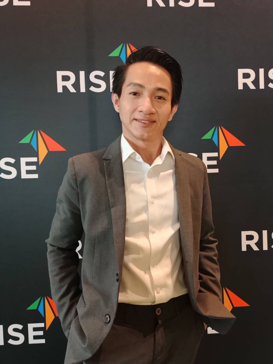 A man in a suit is standing in front of a wall that says rise