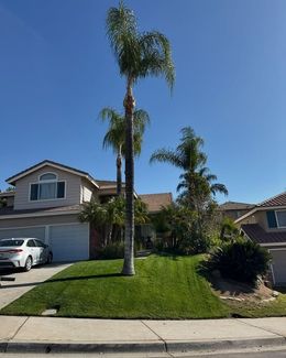 A two-story beige house with palm trees and green lawn on a sunny day. A car is parked in front of the garage.