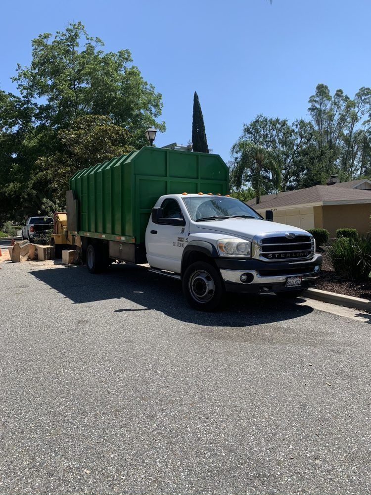 A white and black garbage truck with a green bin is parked on an asphalt road. Trees and a building are in the background.