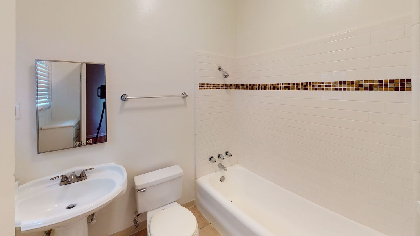 Bathroom with a pedestal sink, toilet, tub, mirror, and towel rack. Off-white walls and tile.