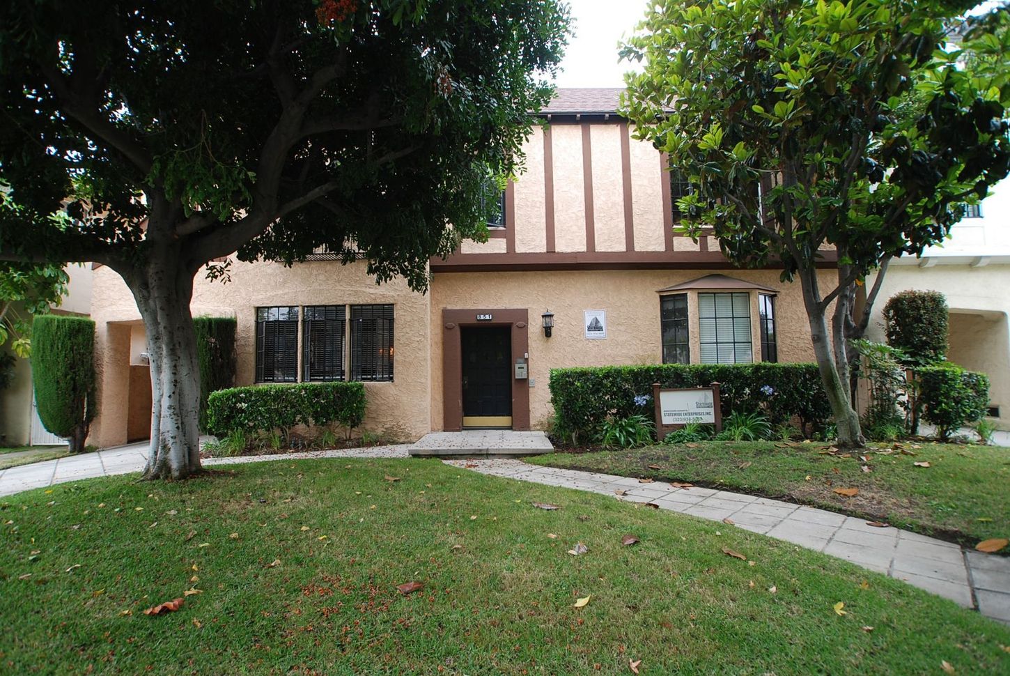 Two-story beige house with brown trim and a green lawn. Trees frame the entrance pathway.