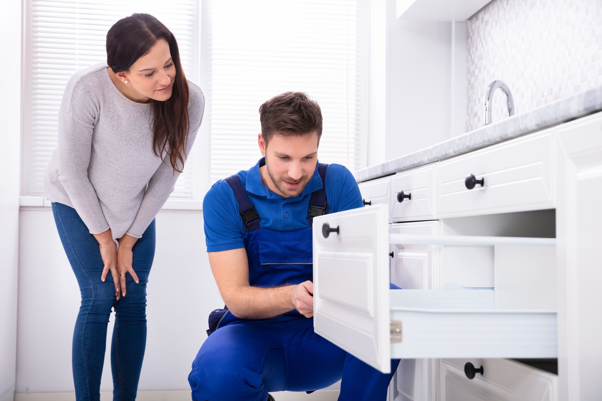 Man in blue jumpsuit fixing a kitchen cabinet drawer as a woman watches. Bright, indoor setting.