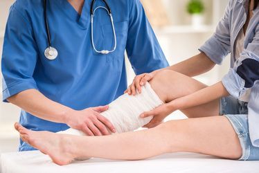 Doctor in blue scrubs examining a bandaged leg of a person in denim shorts.
