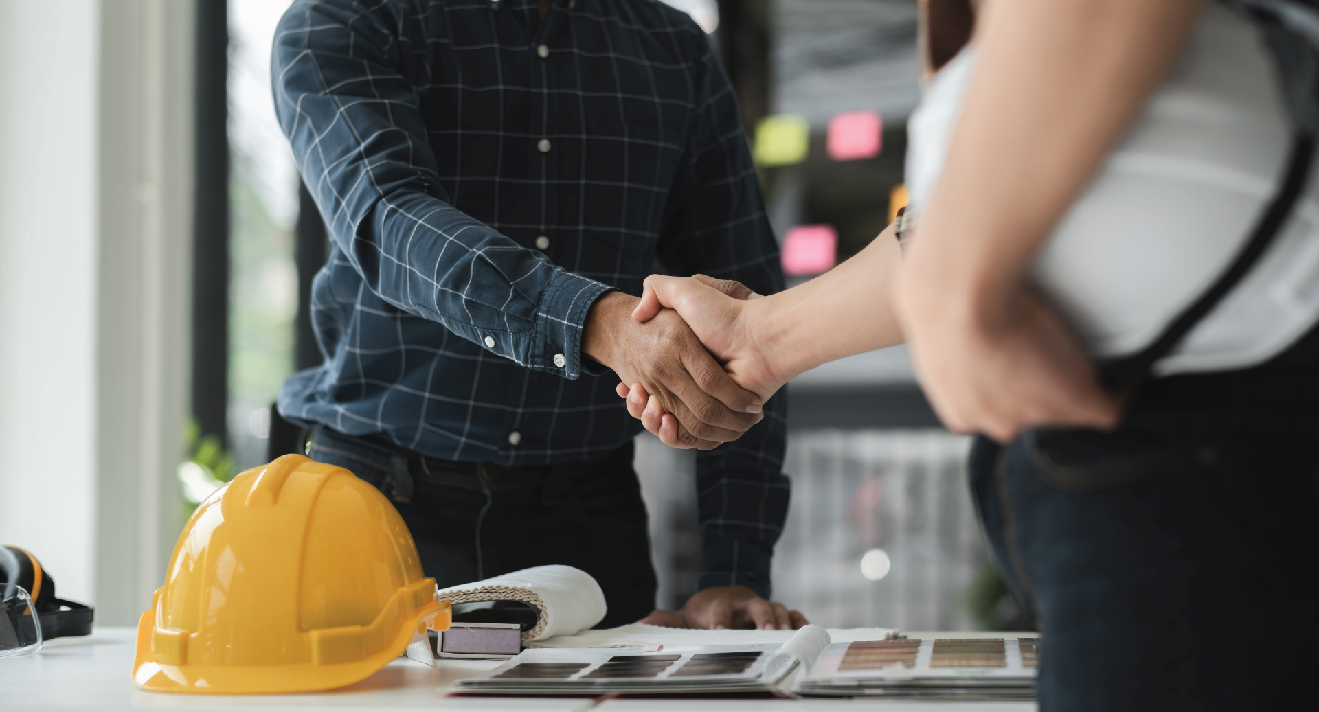 Two people shaking hands over a table with blueprints, a yellow hard hat, and design materials.