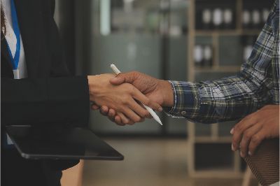 Two people in business attire shake hands, one holding a tablet. Office setting.