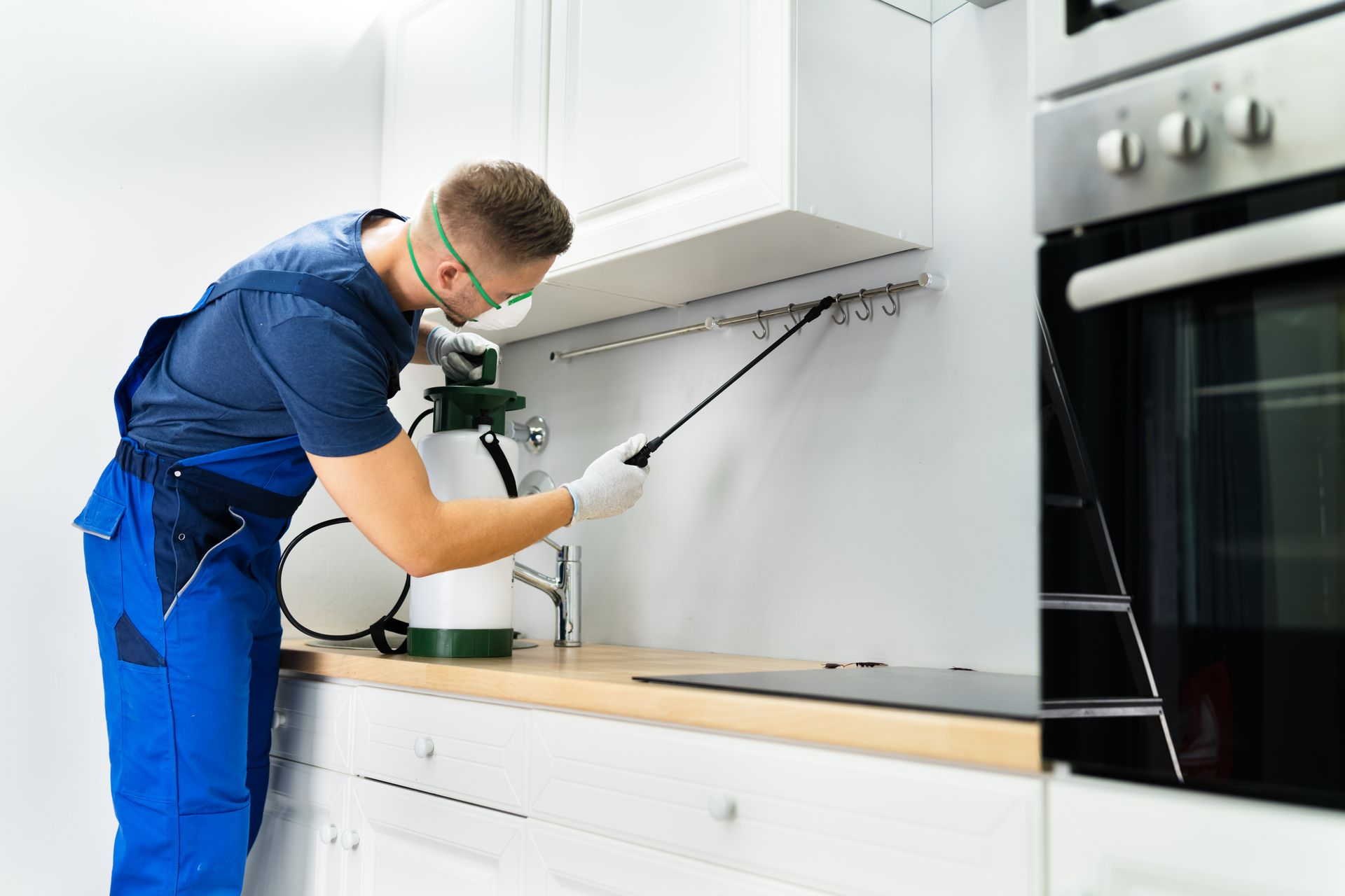 Pest control technician spraying insecticide in a white kitchen, wearing safety glasses and gloves.