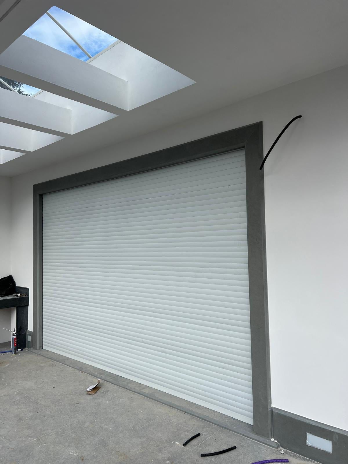 Una serranda di sicurezza avvolgibile di grandi dimensioni e di colore chiaro, installata in una parete bianca con telaio grigio scuro sotto un lucernario.