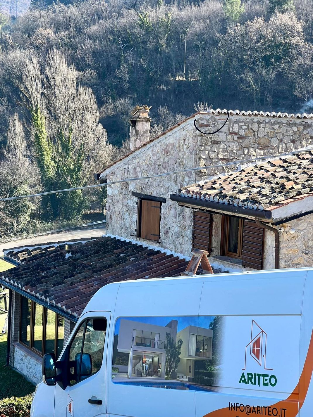 Vista laterale di un furgone bianco parcheggiato davanti a una rustica casa in pietra con tetto di tegole.