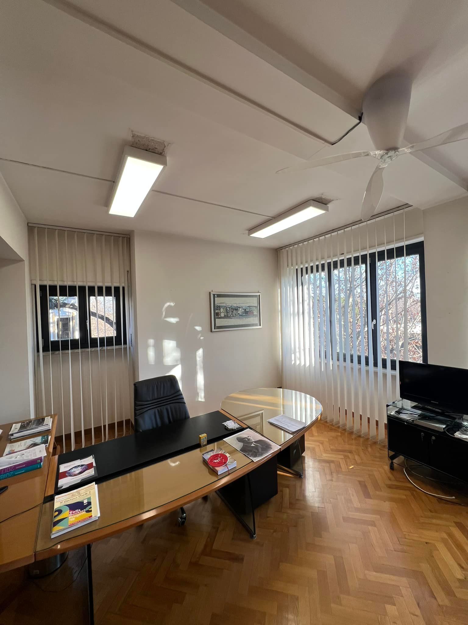 Un ufficio professionale con una scrivania nera a forma di L, una sedia girevole, un televisore e un pavimento in parquet, il tutto illuminato da una luce intensa.