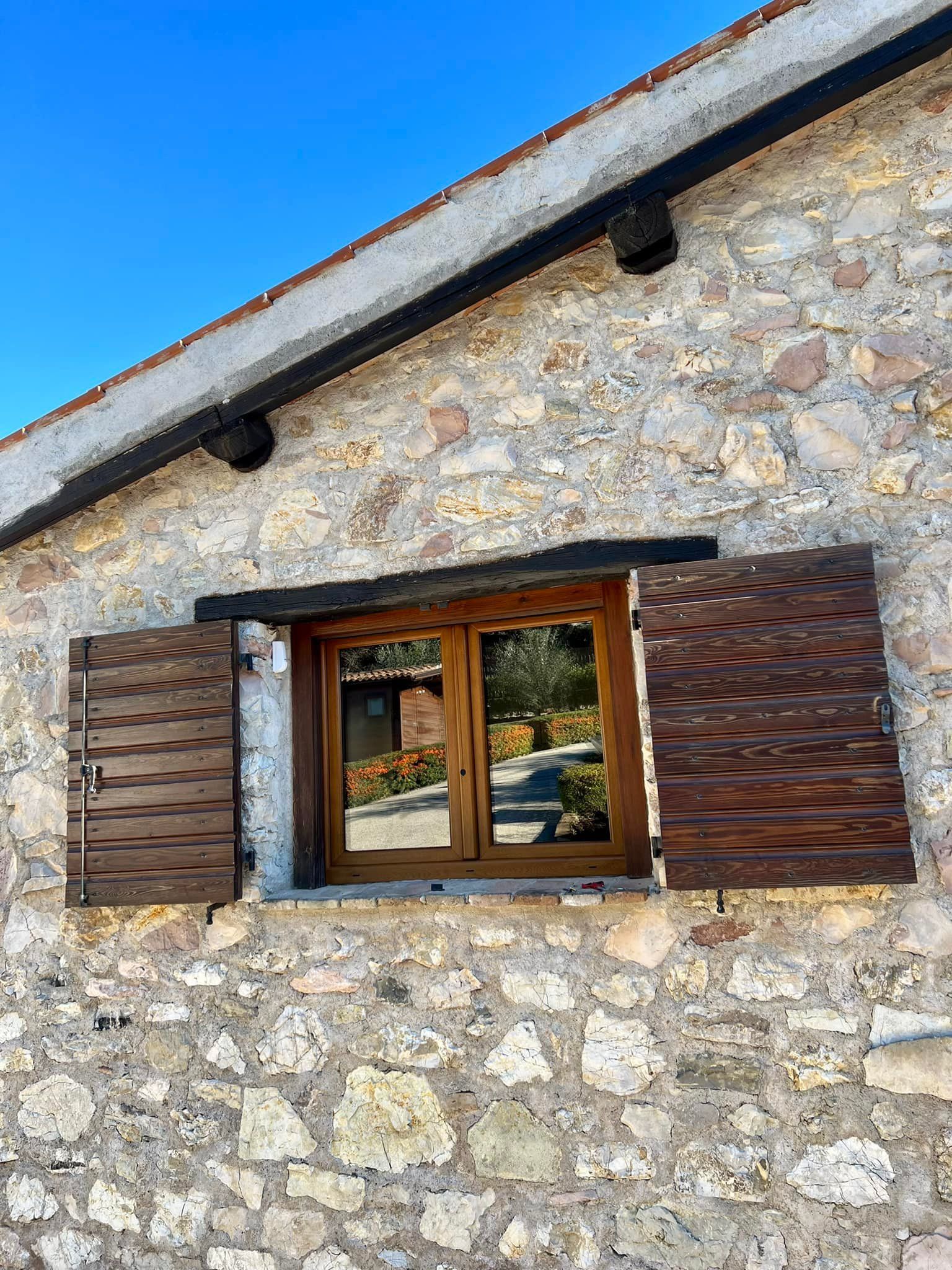 Una finestra con persiane di legno aperte, incastonata in un rustico muro di pietra di un edificio, sotto un cielo azzurro e limpido.