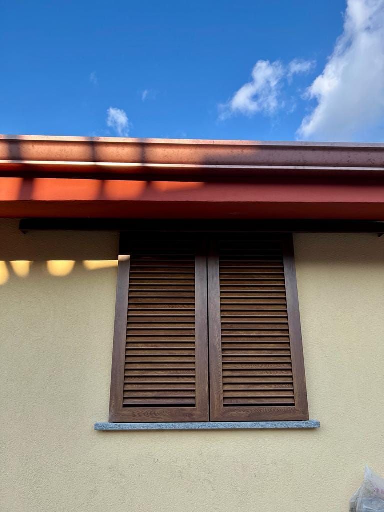 Una finestra con persiane a lamelle marroni su una parete beige, sotto un tetto rosso e un cielo azzurro parzialmente nuvoloso.
