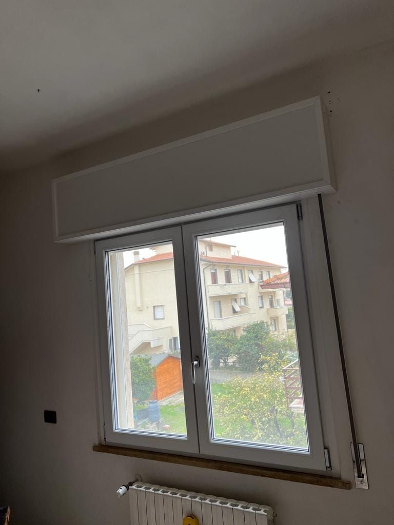 Una finestra bianca a doppio vetro con una serranda avvolgibile chiusa sopra e un termosifone sotto, vista sulle case all'esterno.