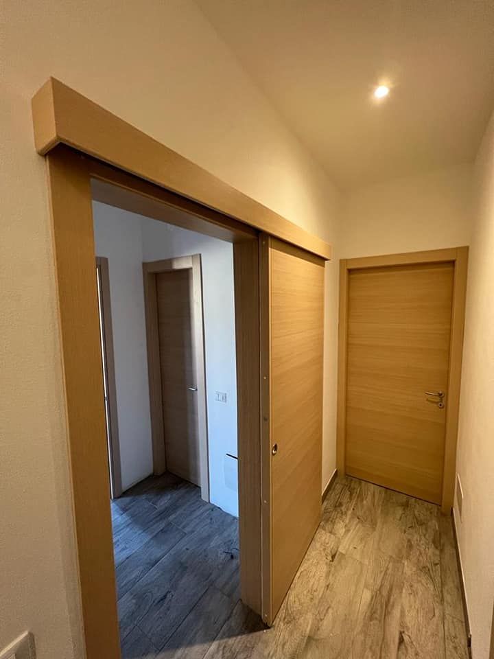 Una porta scorrevole in legno in un corridoio interno dai colori chiari, che conduce a un'altra stanza con una porta e pavimento piastrellato.