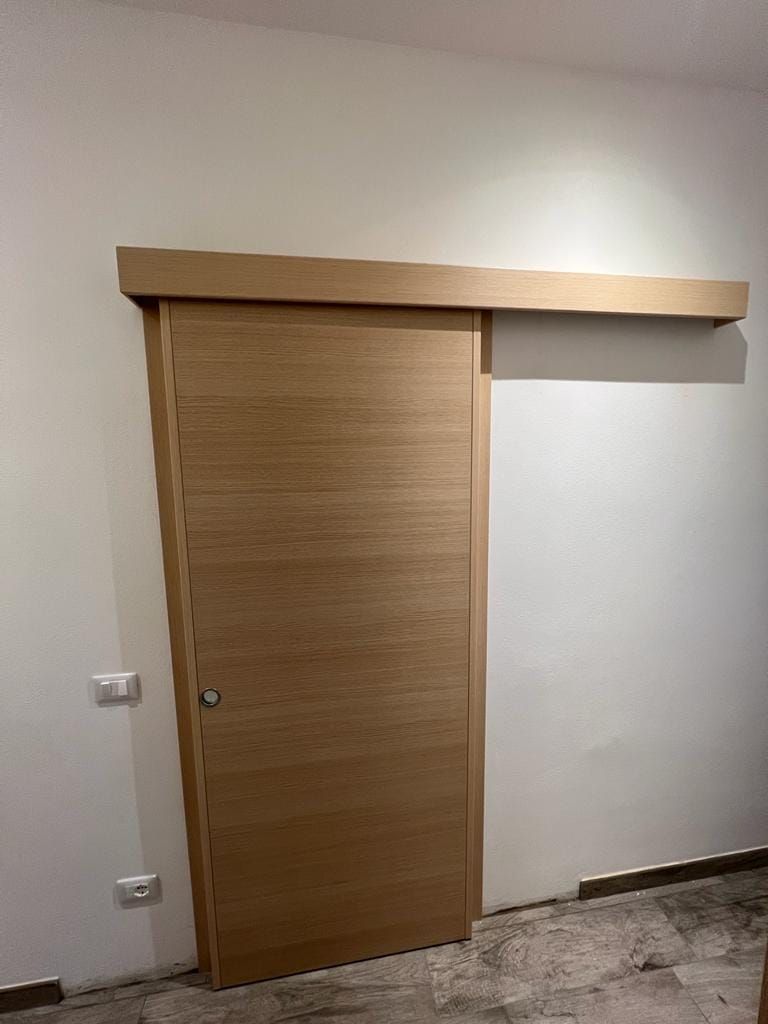 Una porta scorrevole in legno chiaro installata su una parete bianca in un ambiente interno con pavimento in marmo.