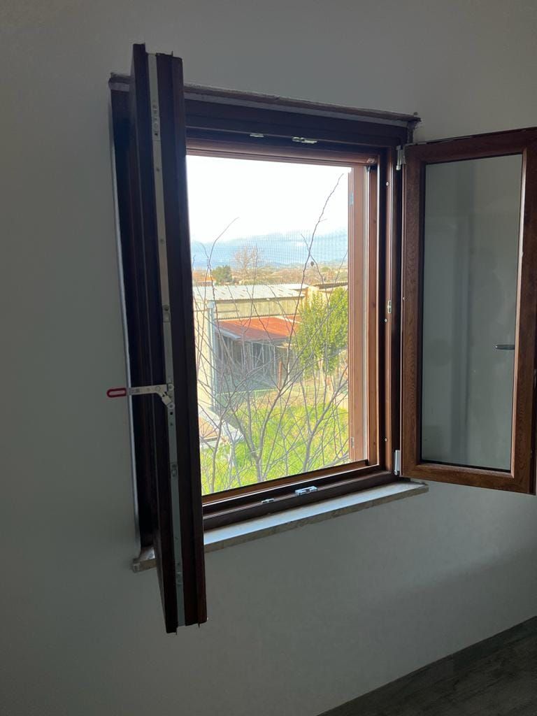 Una finestra con cornice di legno aperta si affaccia su un paesaggio soleggiato con alberi, edifici e colline in lontananza.