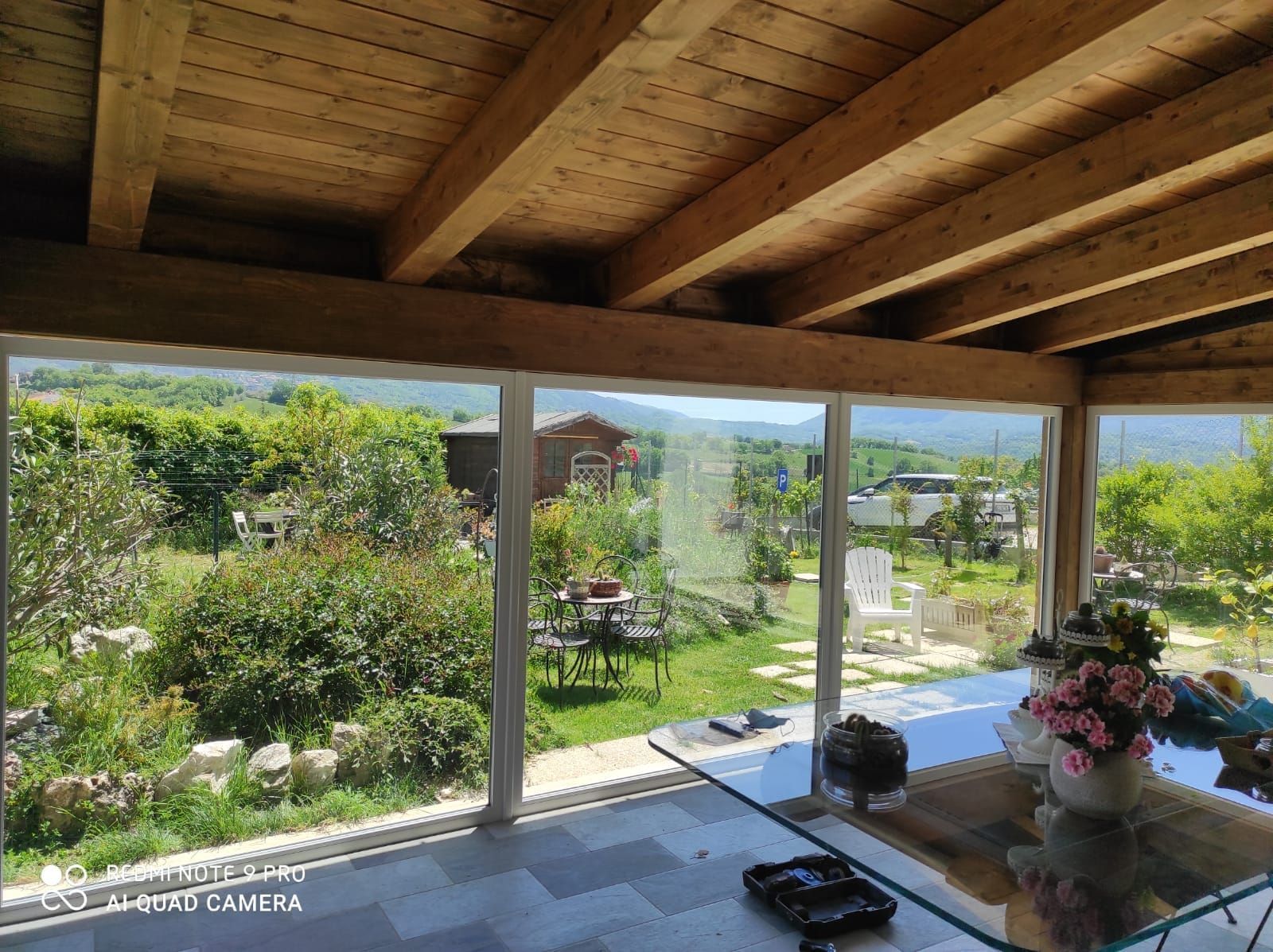 Un patio esterno coperto con un tavolo di vetro, che si affaccia su un rigoglioso giardino verdeggiante e sulle colline in lontananza.