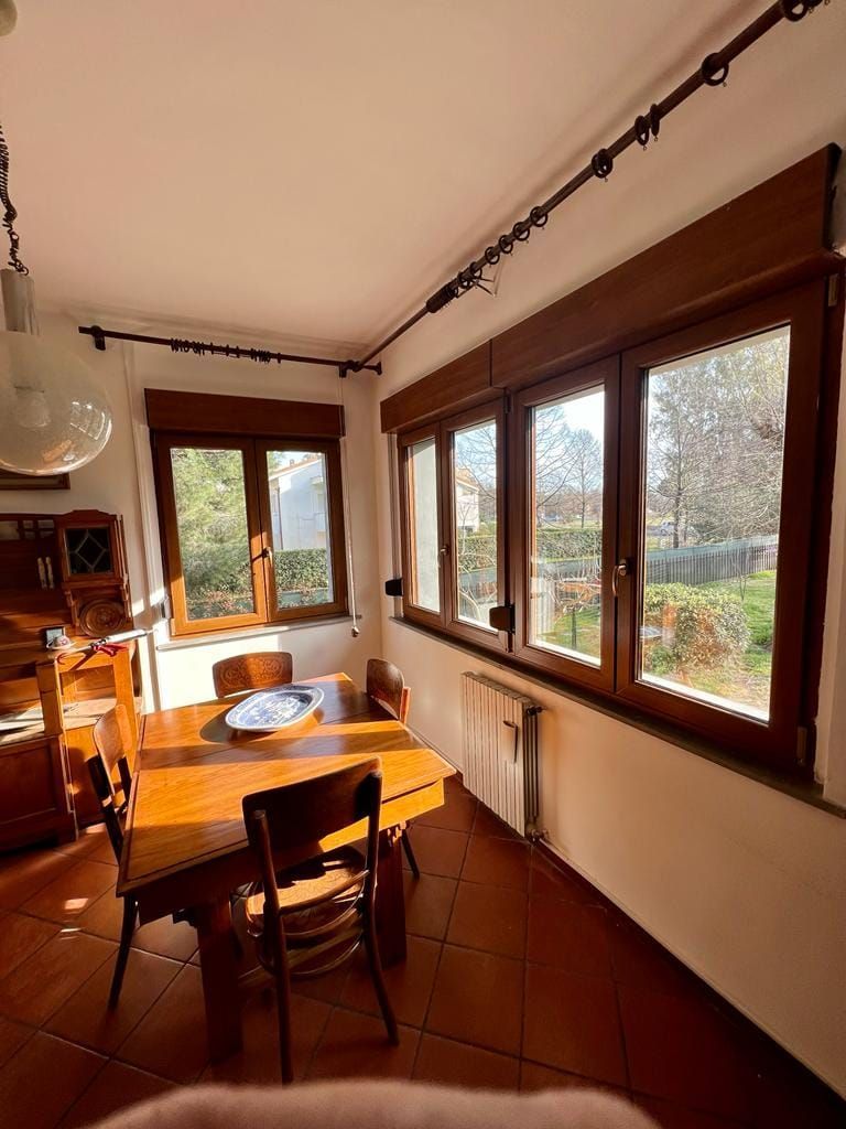 Sala da pranzo con tavolo e sedie in legno e grandi finestre con infissi marroni, inondate dalla calda luce del sole pomeridiano.