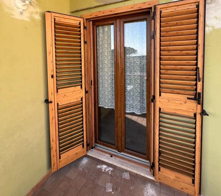 Una coppia di persiane di legno aperte incornicia una porta a vetri con tende bianche su un balcone con pavimento piastrellato.