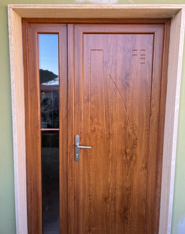 Una porta d'ingresso con finitura effetto legno, pannello laterale verticale in vetro e maniglia in metallo, incastonata in una cornice su una parete verde chiaro.