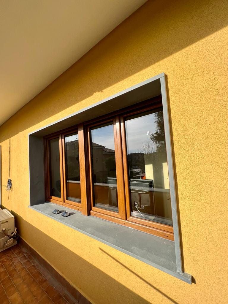 Una finestra con cornice marrone incastonata in una parete esterna gialla e ruvida, con davanzale in pietra grigia.