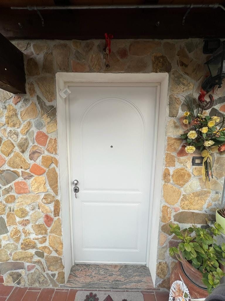 Porta bianca incastonata al centro di un muro di pietra rustica, con una piccola composizione floreale sulla destra e pavimento in piastrelle di terracotta.