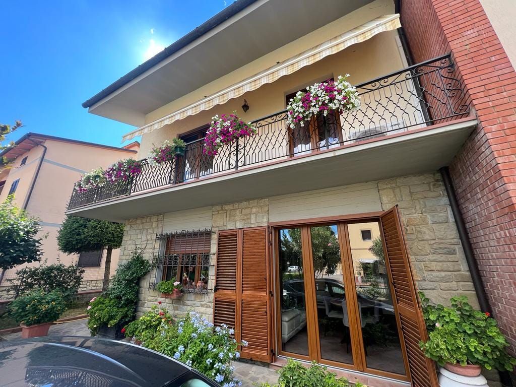 Casa a due piani con un balcone pieno di fiori in fiore, muri in pietra e persiane in legno al piano terra.