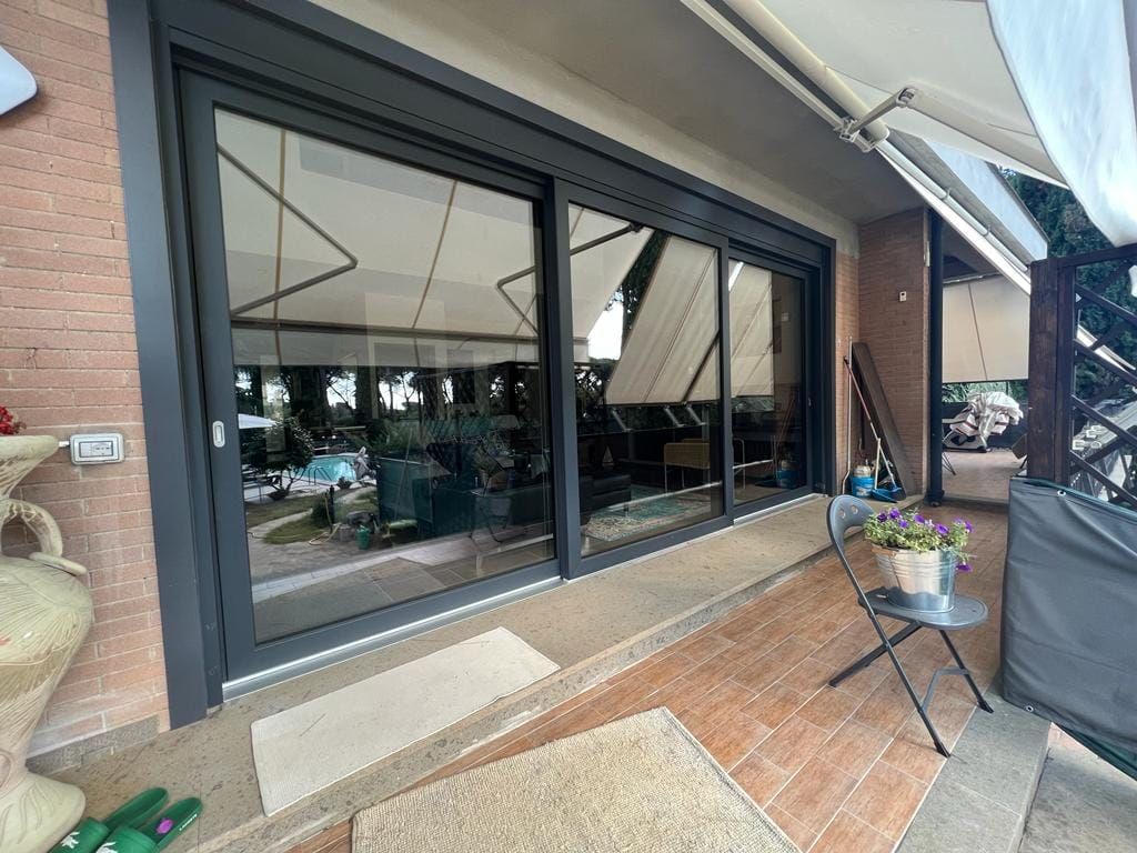 Un patio con ampie porte scorrevoli in vetro, pavimento in mattoni, un tavolino in metallo con una pianta in vaso e una tenda da sole retrattile.