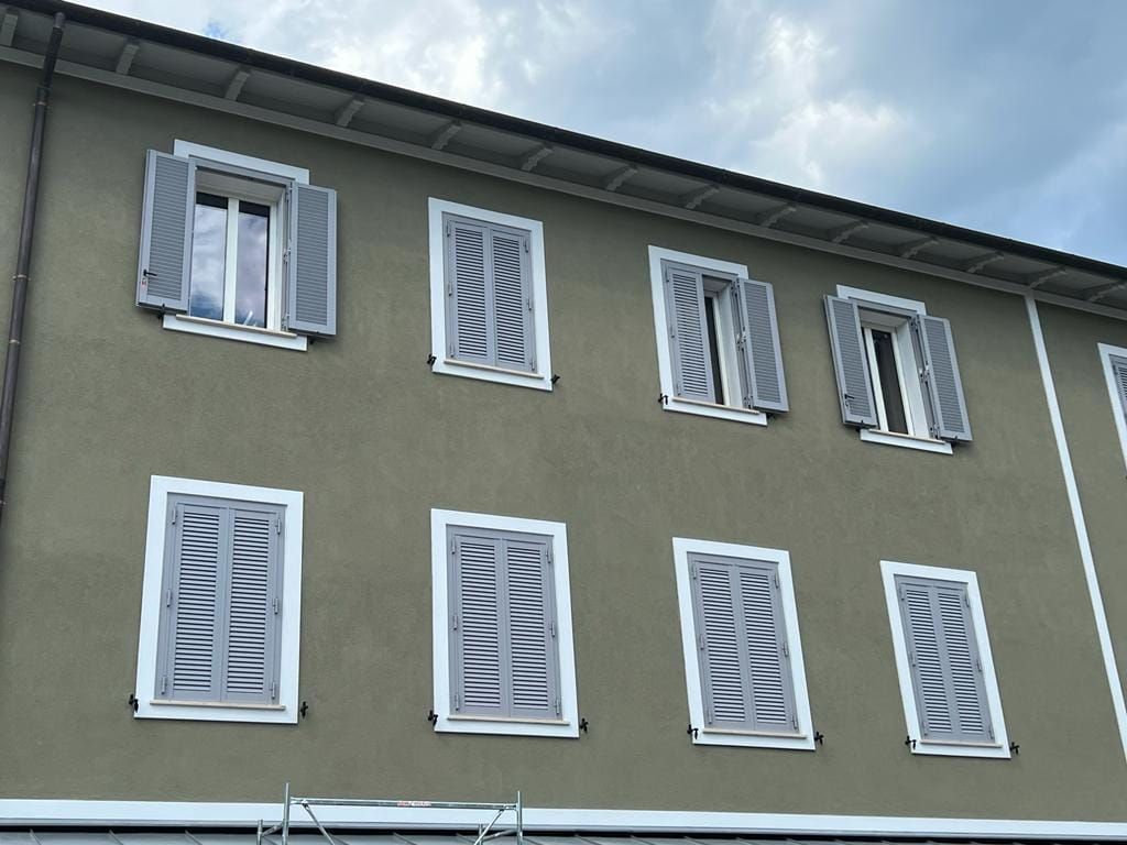 Esterno di un edificio color oliva con due file di finestre dotate di persiane grigie, alcune aperte e altre chiuse.