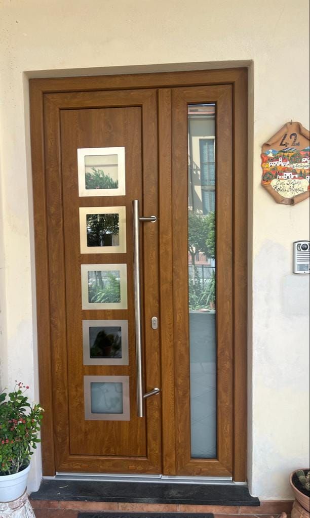 Una porta d'ingresso con finitura effetto legno, maniglia verticale argentata, cinque finestre quadrate e una grande finestra laterale accanto al numero civico.