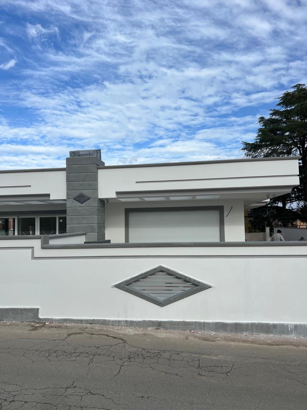 Un edificio basso e moderno, di colore bianco, con un elemento decorativo grigio a forma di diamante sulla parete e sul camino, stagliato contro un cielo azzurro e nuvoloso.